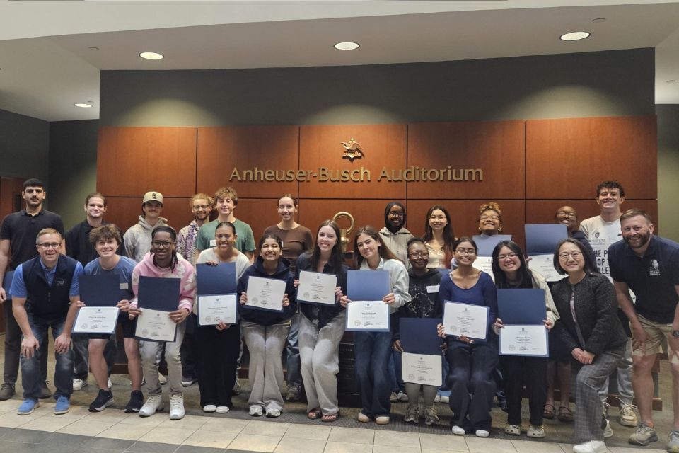 a large group of people stand posed holding certificates in front of the Anheuser Busch Auditorium