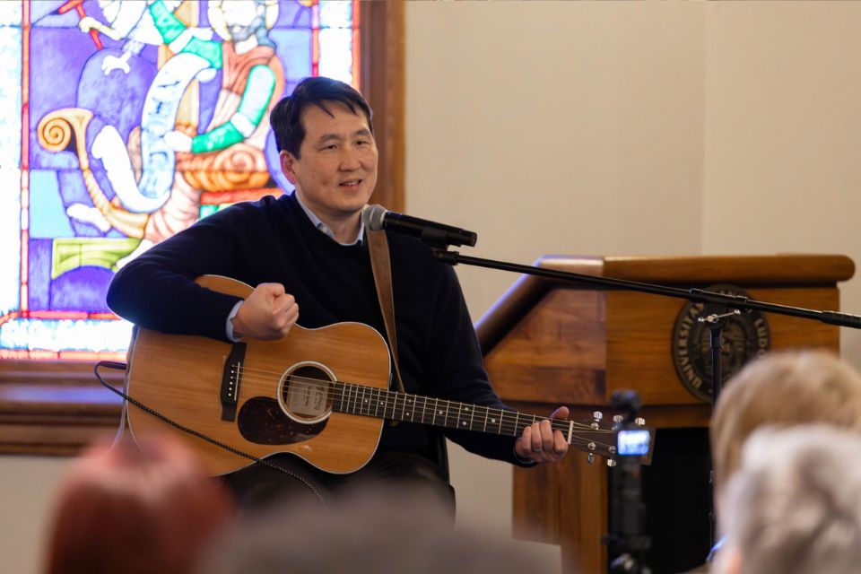 a man sits with a guitar speaking in front of a crowd of people
