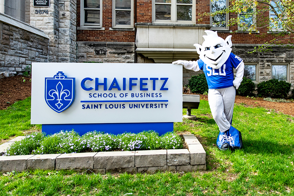 The Saint Louis Billiken mascot poses next to the Chaifetz School of Business sign outside of Cook Hall