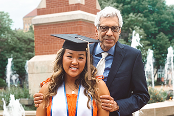 One-Year MBA graduate Cassidy Manns poses for a photo with her gap and honors regalia in front of the Clock Tower fountains with SLU President Fred Pestello, Ph.D. 