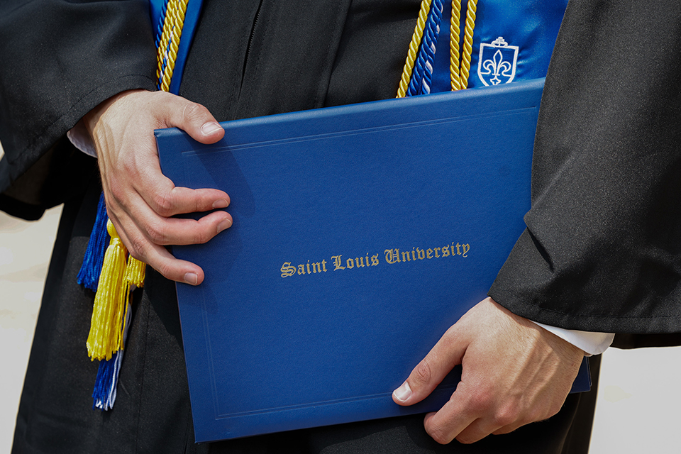 A student in graduation robes holds a Saint Louis University diploma frame