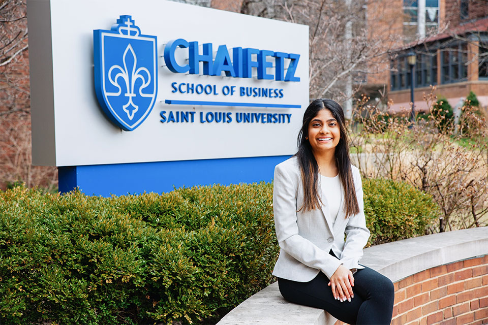 Nisha Vodrahalli , wearing a suit, sits in front of a sign for the Chaifetz School of Business.