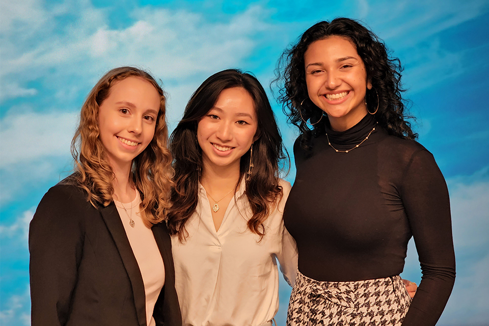 Darcy Taylor, Elizabeth Wangley, and Athena Valera-Barrios posed looking at the camera in front of a backdrop with clouds and blue sky