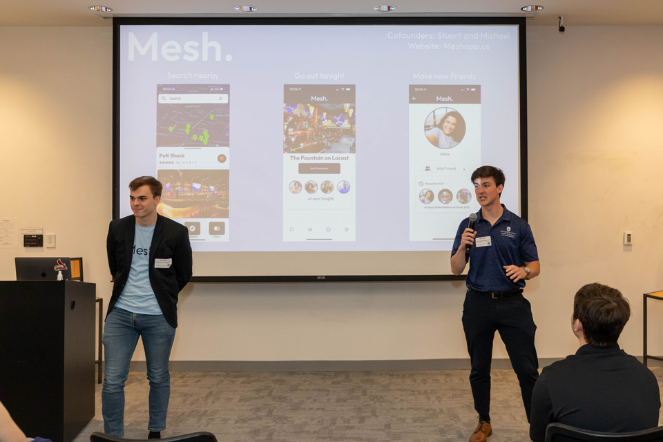 Two students stand at the front of a classroom, in front of a whiteboard with a presentation for their Mesh app.