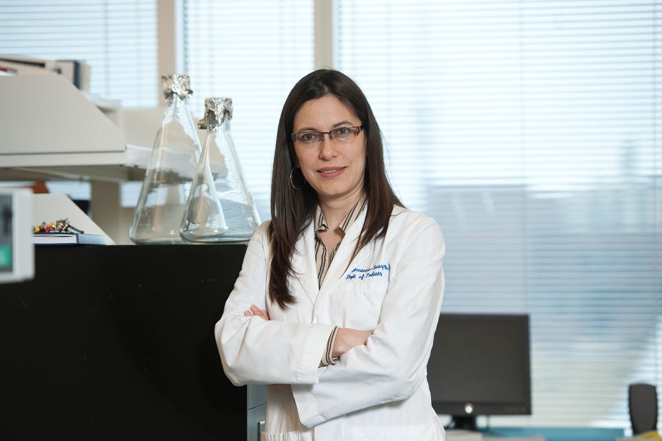 Wearing a white lab coat, Adriana Montano, Ph.D. looks at the camera while standing in her lab.