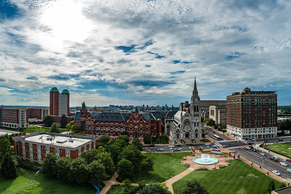 SLU's north campus as seen from an aerial view looking east, with a blue sky and backlit clouds.