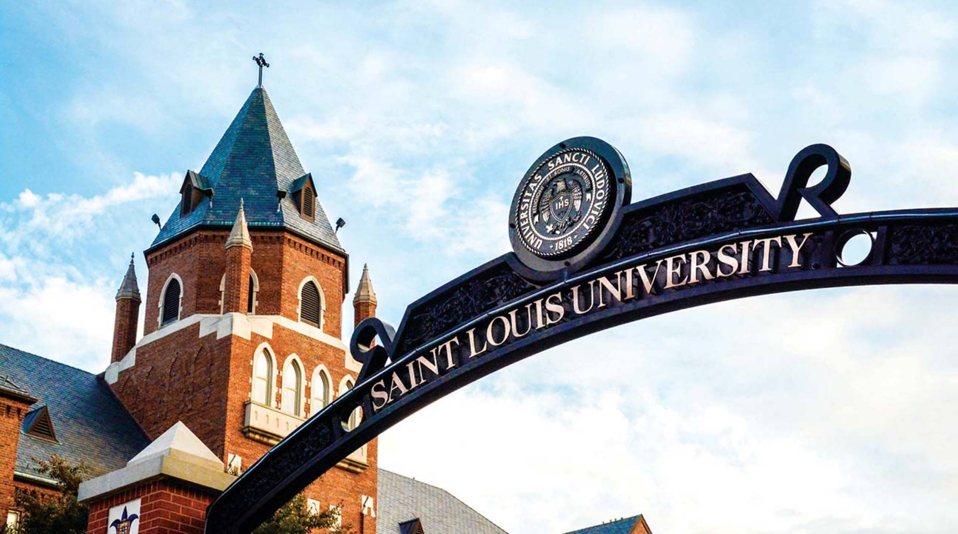 Looking up at an arched entrance gate on SLU's north campus, with the spire of Cook Hall and blue sky in the background.