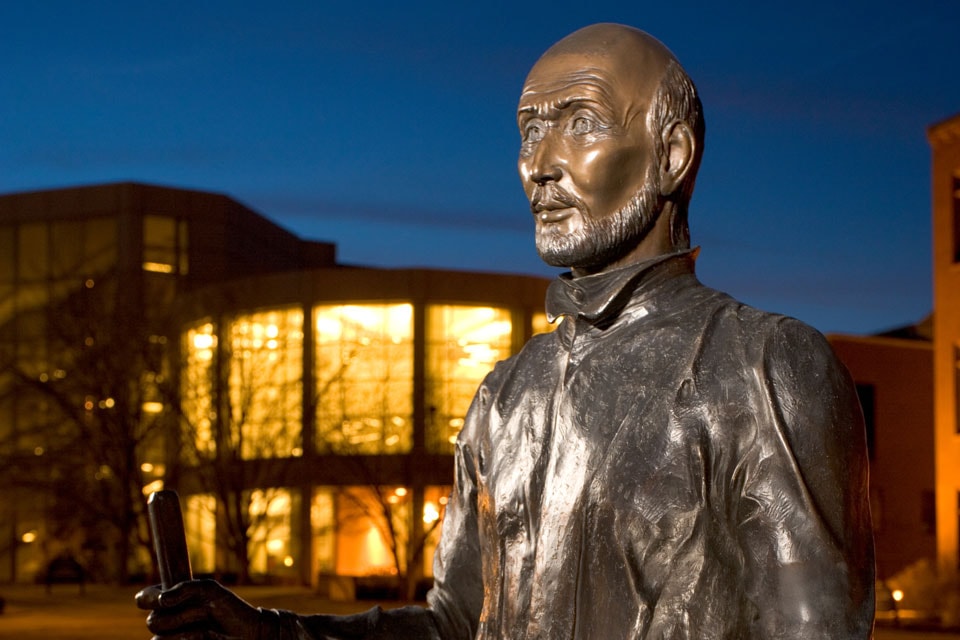 The face and shoulders of a bronze statue of St. Ignatius at dusk, with SLU's Pius X Library in the background.