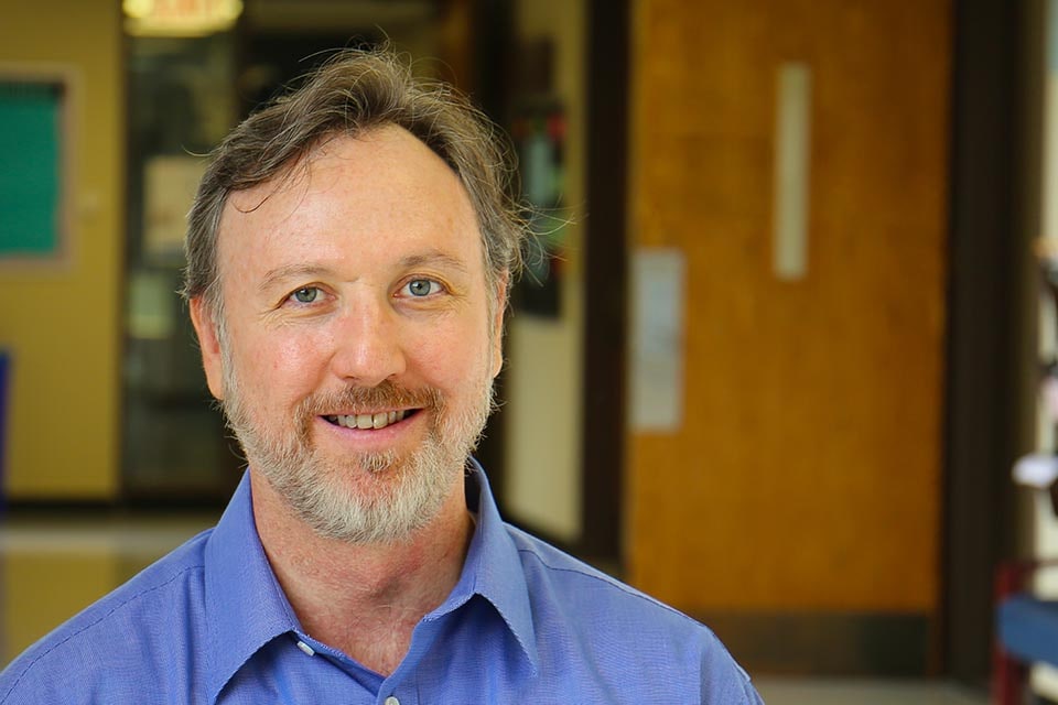 Michael Vaughn smiles at the camera while standing in the hallway of an academic building.