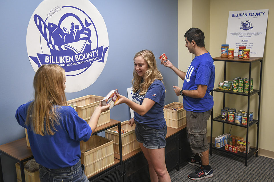 Three students examine packages of food amongst baskets and shelves of donated goods.