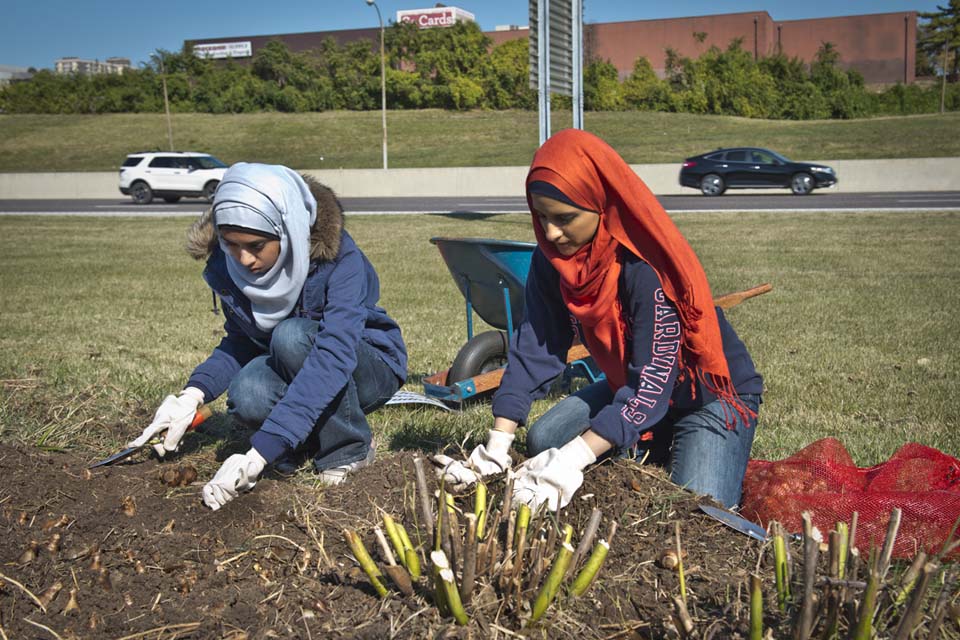 Two SLU students kneel next to a gardening plot, wearing gloves, headscarves and digging in the dirt.