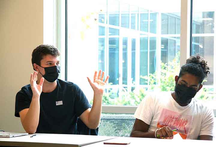 A student in a mask with both hands raised sits next to another student at a classroom table