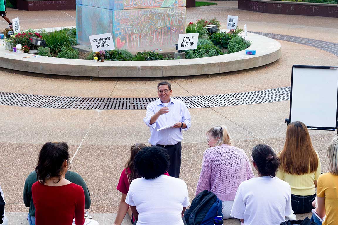 A professor stands at the center of an outdoor lecture space with rows of students seen from the back