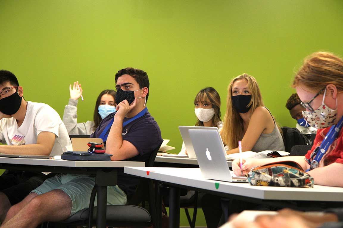 Students in masks sitting in a classroom listening to a lecture; one student raises her hand.