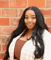 Aubra Ladd Headshot Aubra Ladd, headshot, stands in front of a brick wall and smiles. She is wearing a tan and white plaid shirt over a brown top.