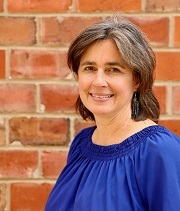 Leah Sweetman Headshot Leah Sweetman, headshot, wears a blue blouse and stands in front of a red brick wall, smiling.