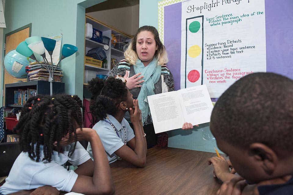 A teacher holds a book open while elementary students sit at a table in a classroom. A bulletin board behind the teacher reads "stoplight paragraphs."