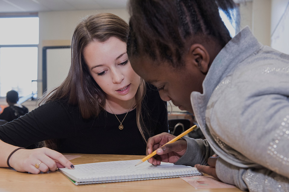 Two students look at a notebook while one hold a pencil.