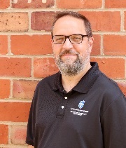 Bryan Sokol, headshot, wearing a black SLU polo shirt, stands in front of a brick wall smiling.
