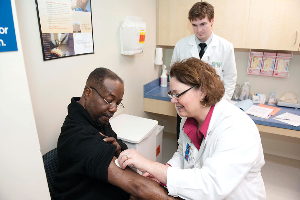 A health care worker holds something against a patient's arm while another health care worker observes.
