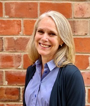Melissa Apprill, headshot, wearing a navy blue sweater and light blue blouse, stands in front of a brick wall and smiles.