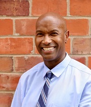 Michael Jackson, headshot, wearing a blue button-down shirt and blue striped tie, stands in front of a brick wall and smiles.