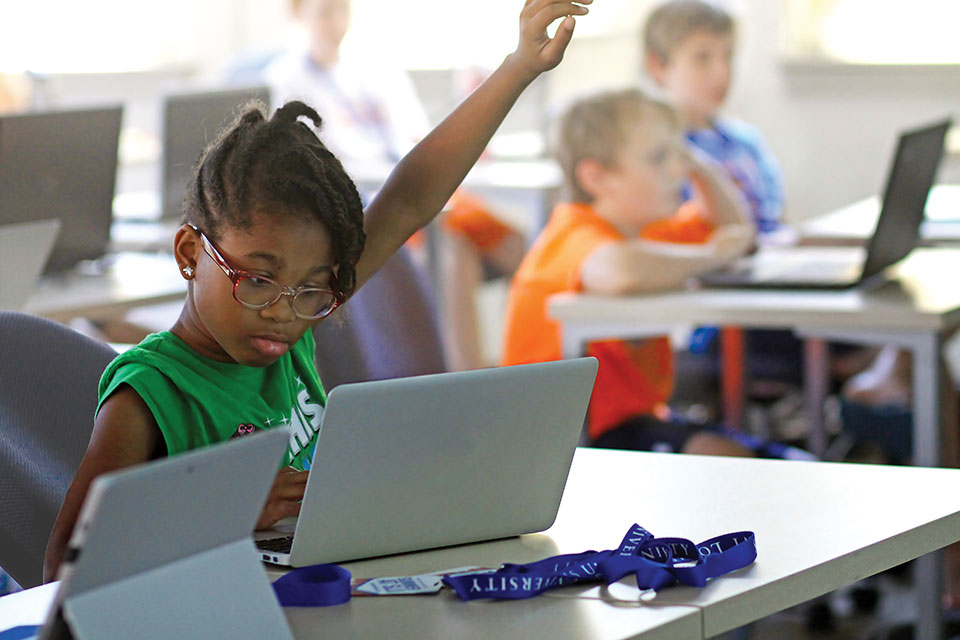 A child looks at a laptop screen and raises her hand while sitting in a classroom with other students her age.