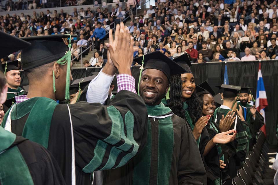 Two students wearing caps and gowns high five each other in a crowded arena.