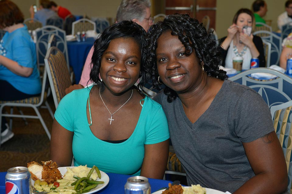 Two students pose while sitting at a dinner with plates of food and soda cans in front of them. 