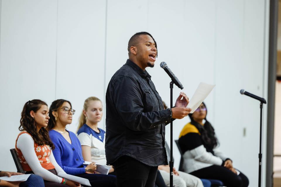 A student stands and speaks into a microphone holding a piece of paper while other students, seated, look on