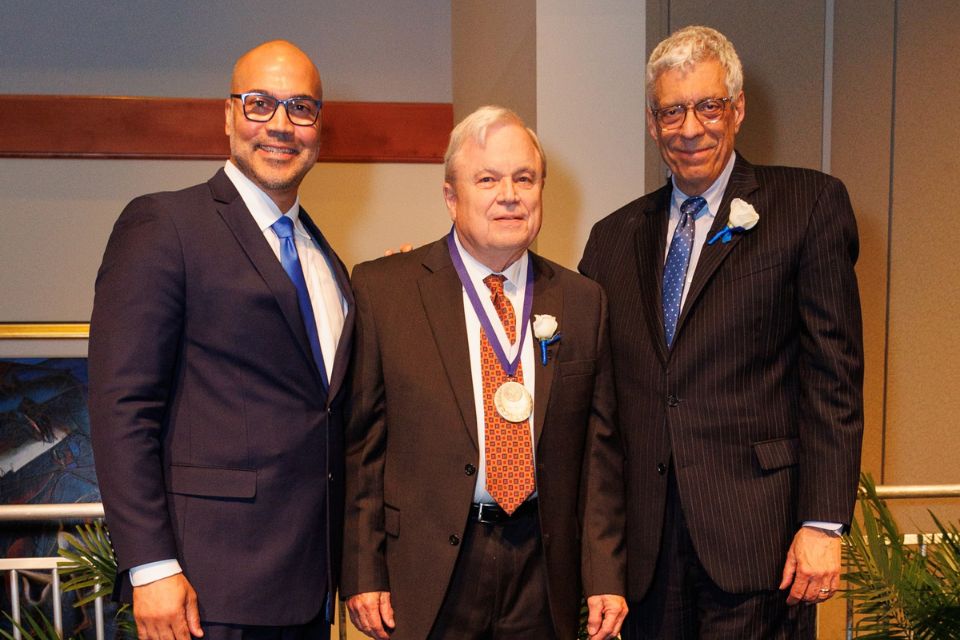 Three men in suits stand on the stage during an award ceremony. The one in the middle wears a medal.