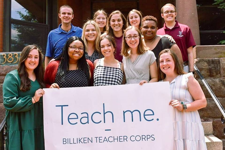 A group of 12 teachers corps members stand on steps outdoors while holding a sign that reads "Teach me. Billiken Teachers Corps."