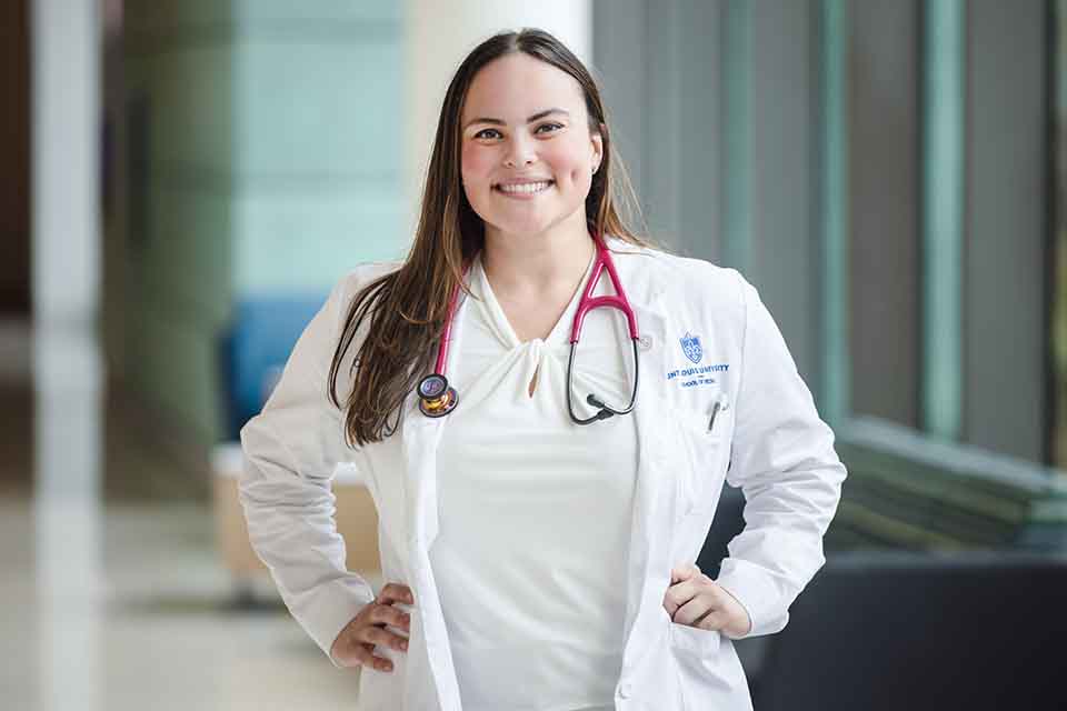 Caroline Wright standing in a lab coat with a stethascope in a lounge