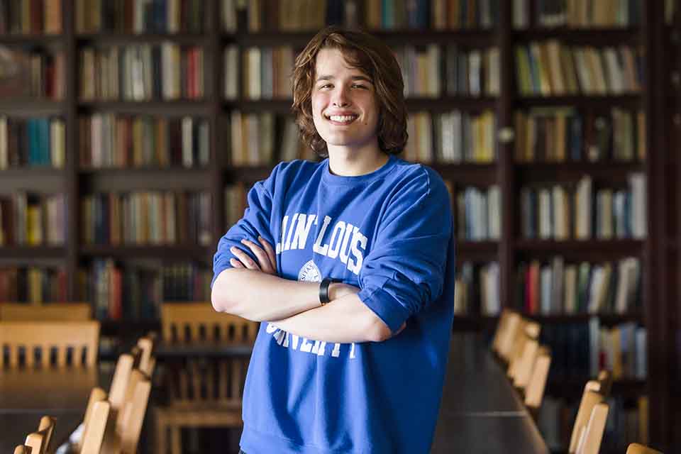Sam Huster in a SLU sweatshirt standing in front of a row of bookcases