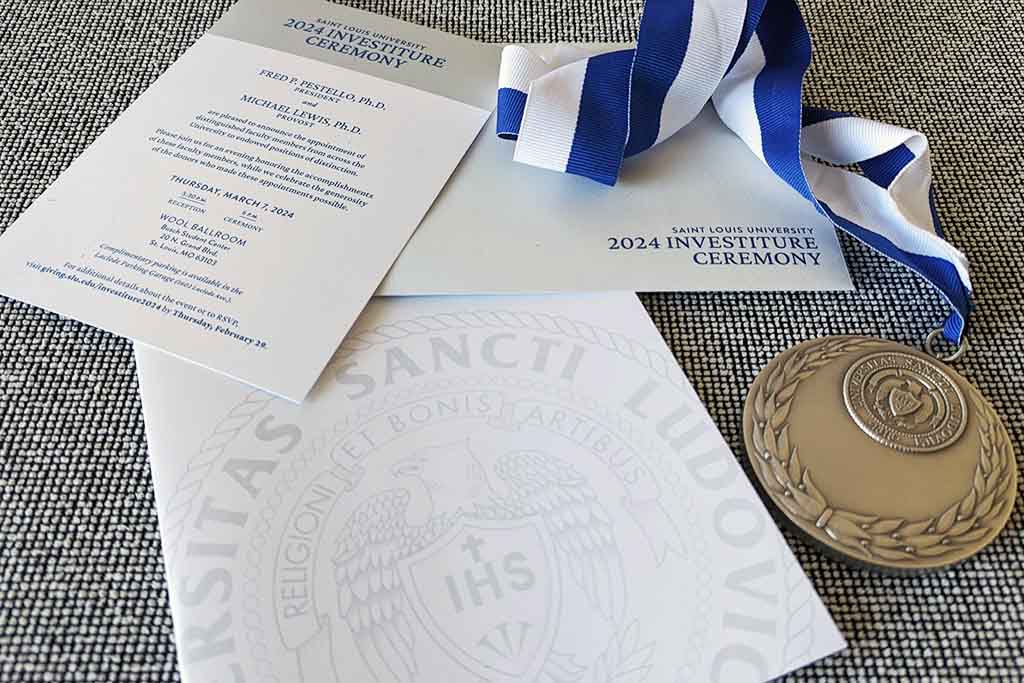 The invitation, envelope and program for SLU's 2024 investiture ceremony laid out with a medallion that will be presented to honorees on a grey tweed background.