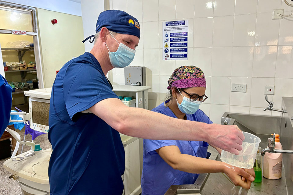 A man pours water over a womans hands. They both wear scrubs and masks.