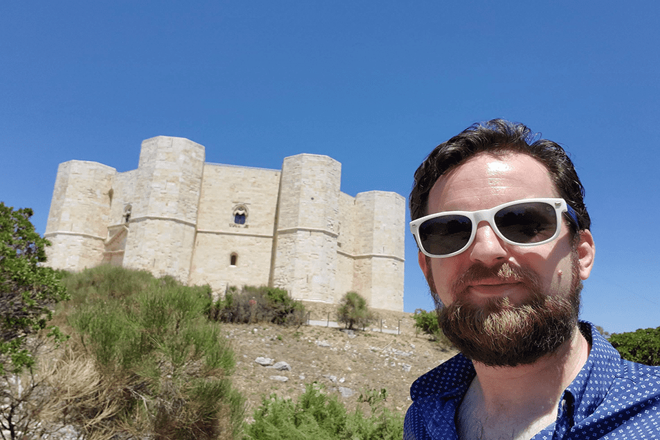 Thomas Morin, wearing sunglasses, looks at the camera with a medieval castle in the background.