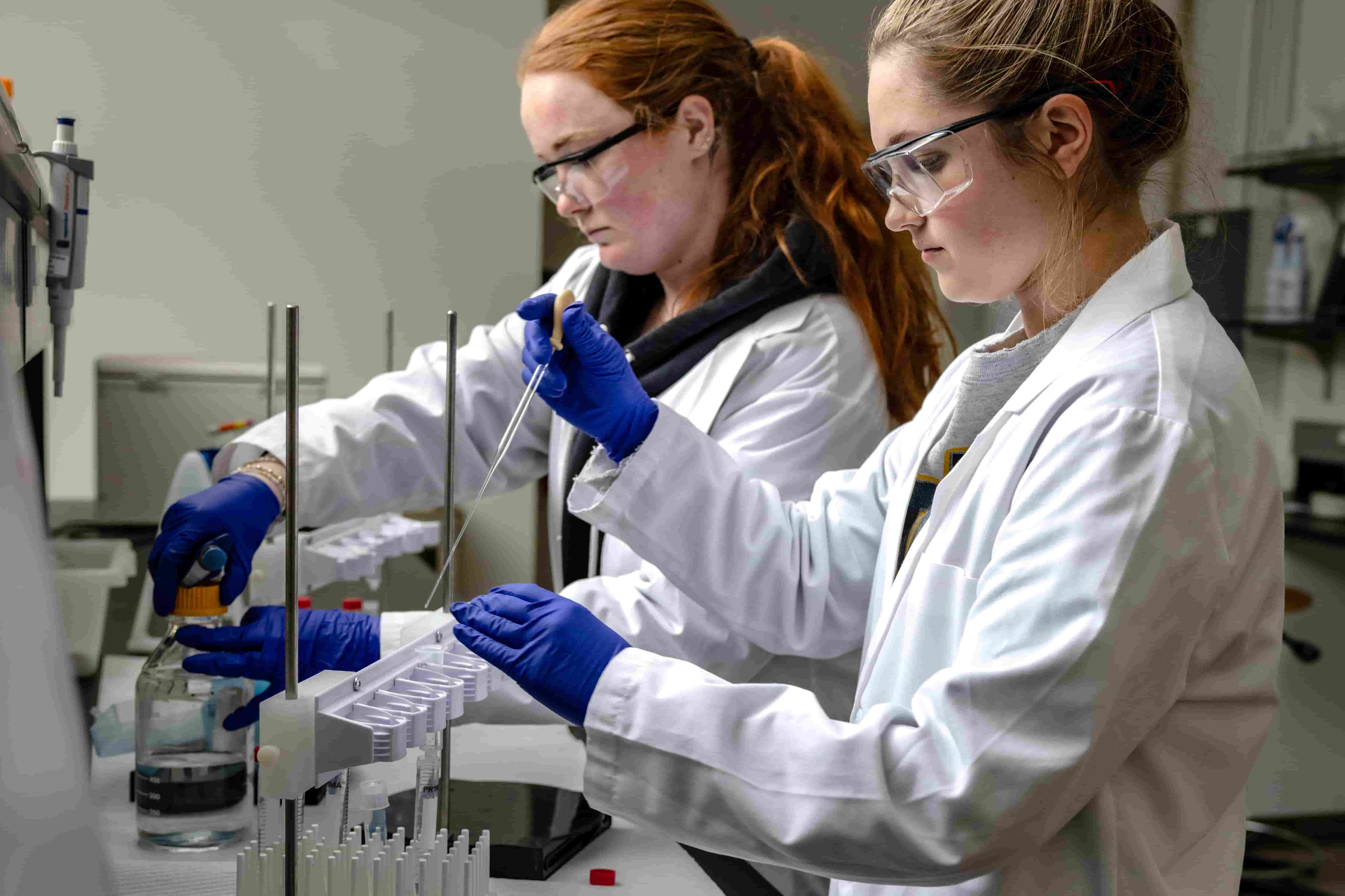 Two students wearing lab coats, gloves and safety goggles conduct an experiment together in a science laboratory.