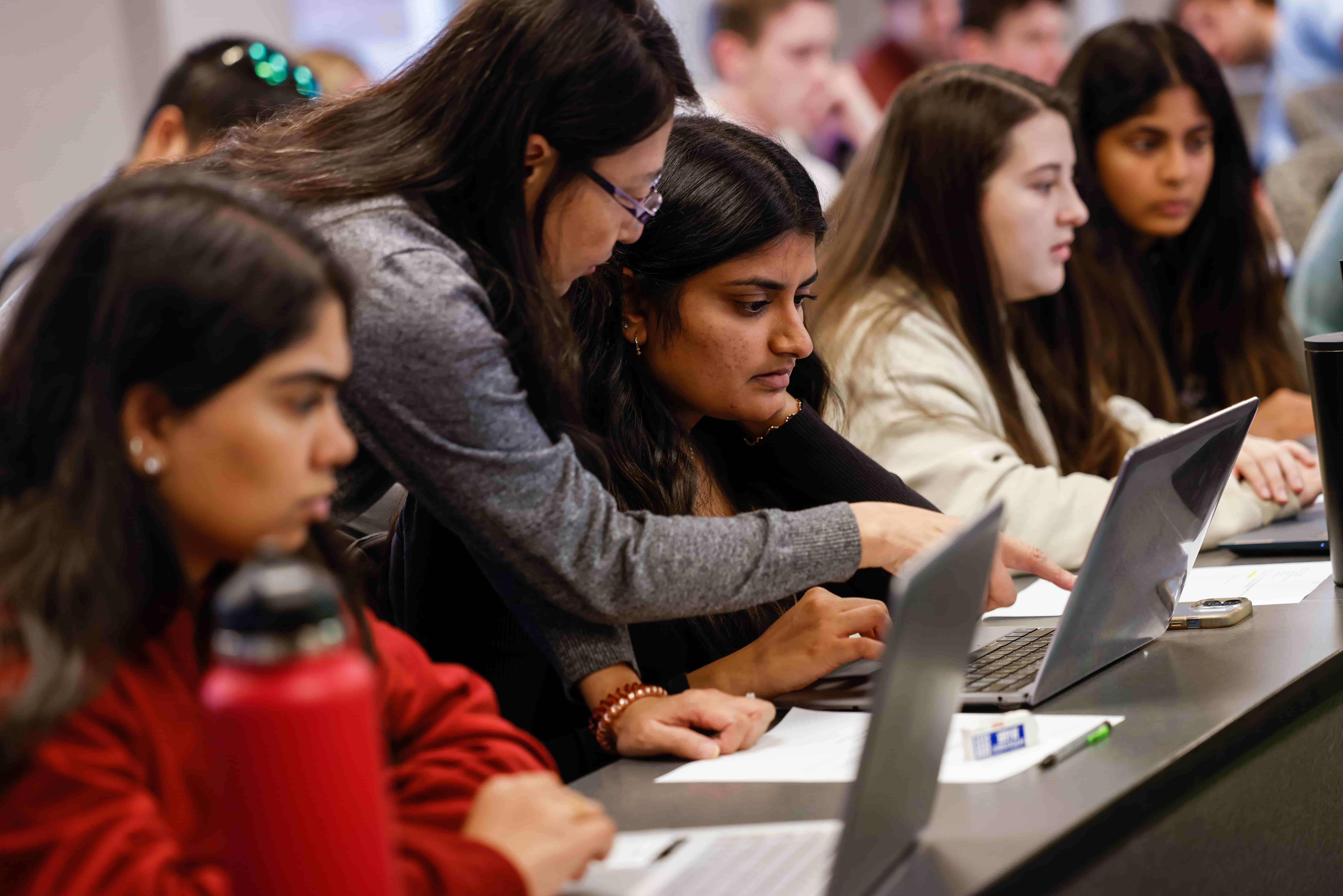 A professor assists a student on her laptop while other students work nearby in a campus classroom.