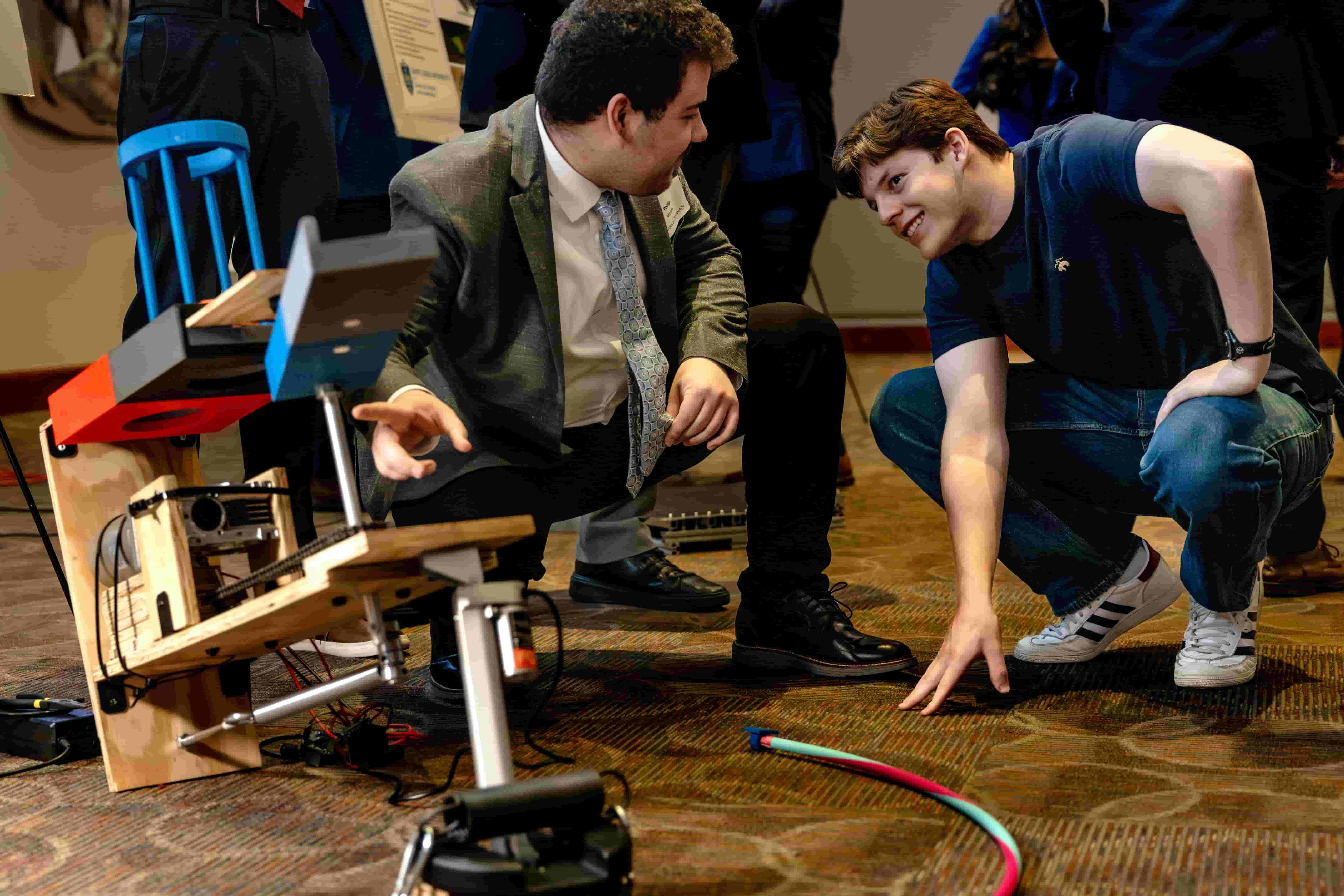 Two people crouch beside a mechanical project and discuss its design during a campus symposium.