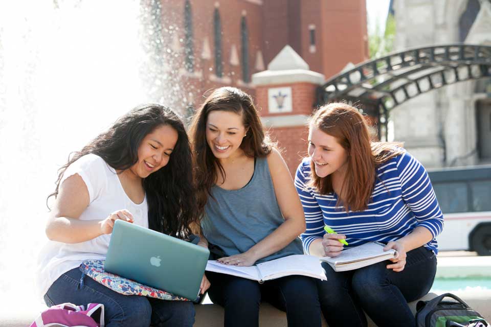 Three students share a look at a laptop, with notebooks also on their laps, while sitting in front of a fountain.