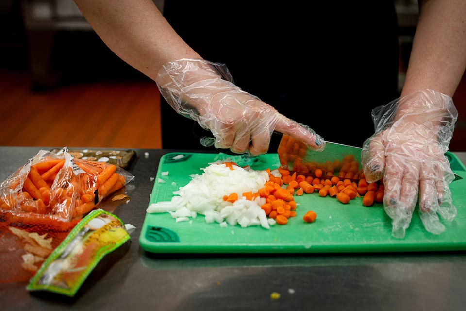 A person seen from the torso only in an apron with hands with a knife chopping carrots on a cutting board