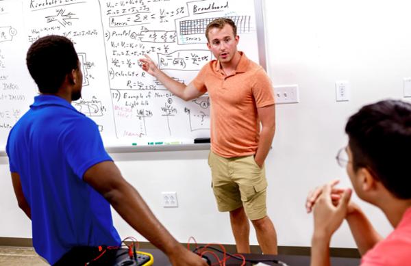 A student at whiteboard gestures to a formula while two other students look on