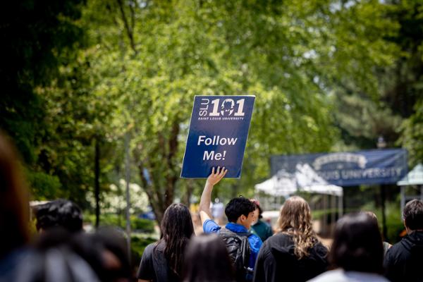 A SLU 101 leader seen from behind in a crowd gives a tour to new students holding a sign that says Follow Me