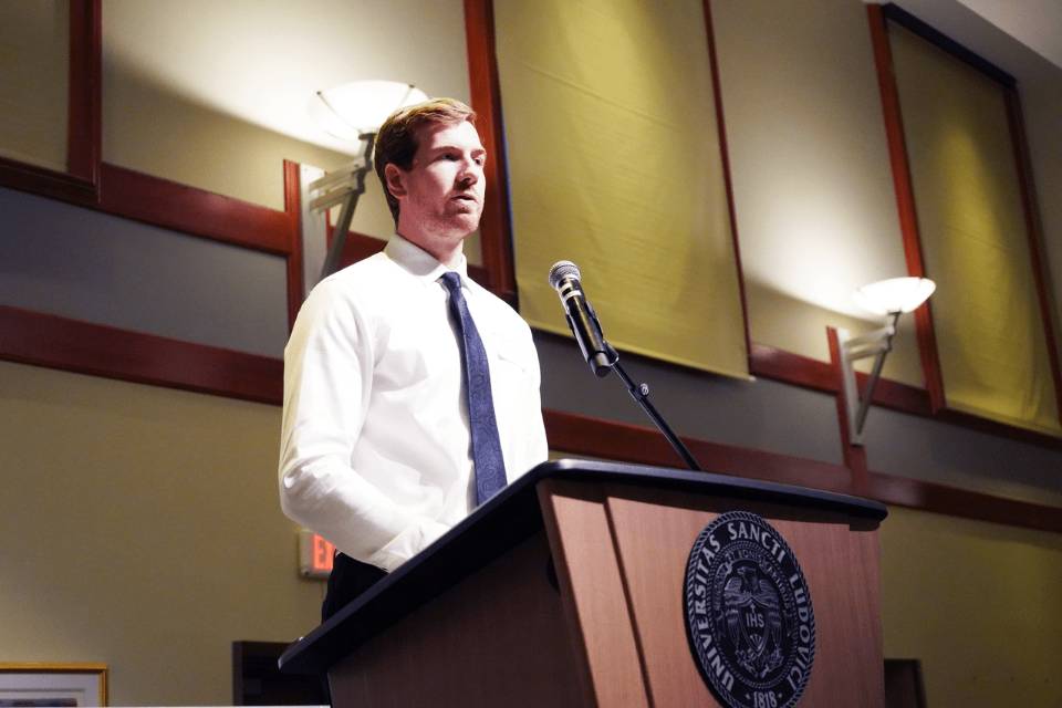 A student stands talking at a lectern that has the SLU seal on the front of it.