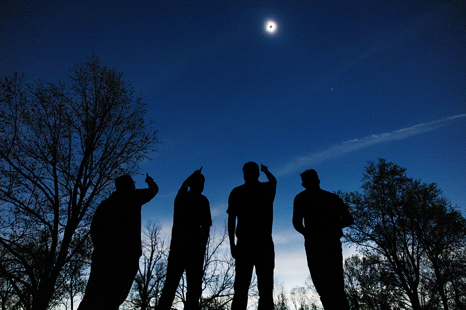 Four students are seen from behind, under a dark sky, with light increasing toward the ground. They are looking up and pointing at the sun with the moon passing in front of it.