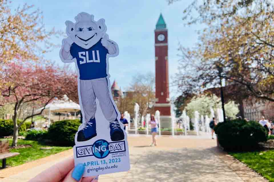 A hand holding a paper cutout of the SLU Billiken in front of the Clock Tower with the words Giving Day April 13, 2023 givingday.slu.edu