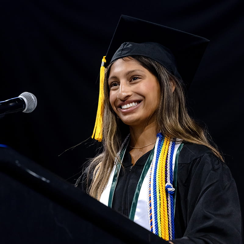 A graduate wearing cap and gown smiles while standing in front of a microphone.