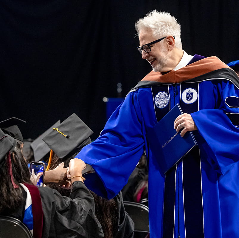 James Gunn in SLU academic regalia robe, holding a diploma shakes hands with graduating students in commencement gowns and hats.