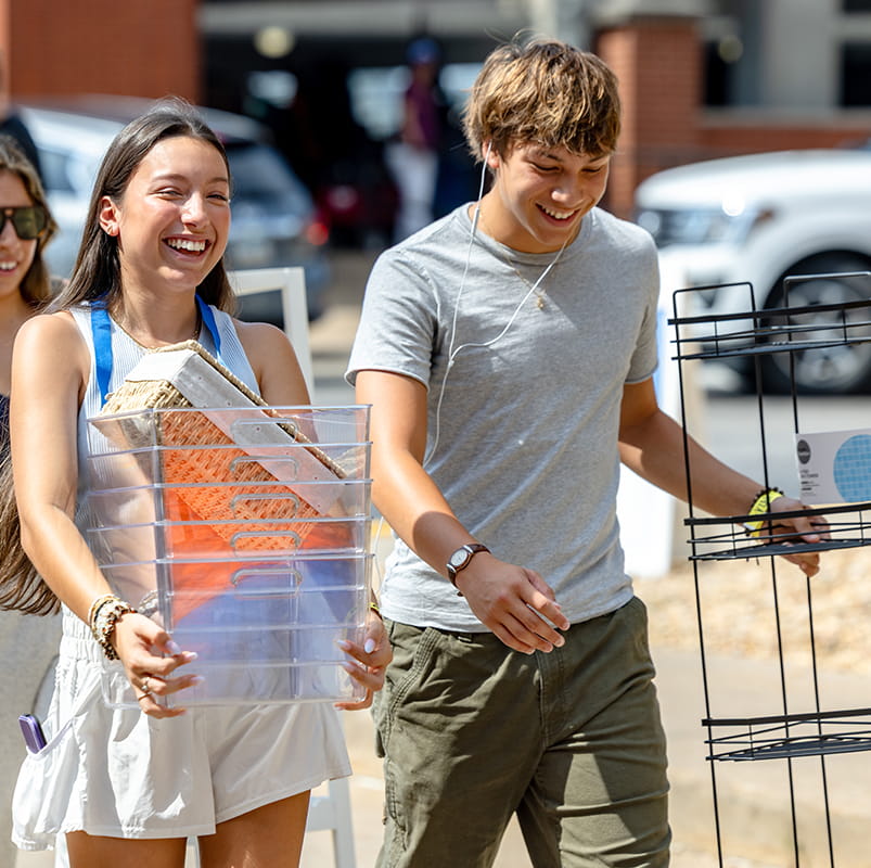 Two students walk toward a dorm on move in day. One carries plastic storage bins and the other carries a metal shelf set.